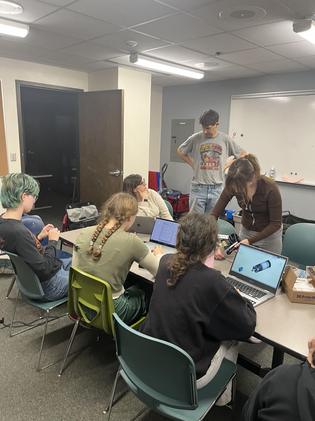 Teammates working on laptops, CAD, and robot parts around a table with a mentor